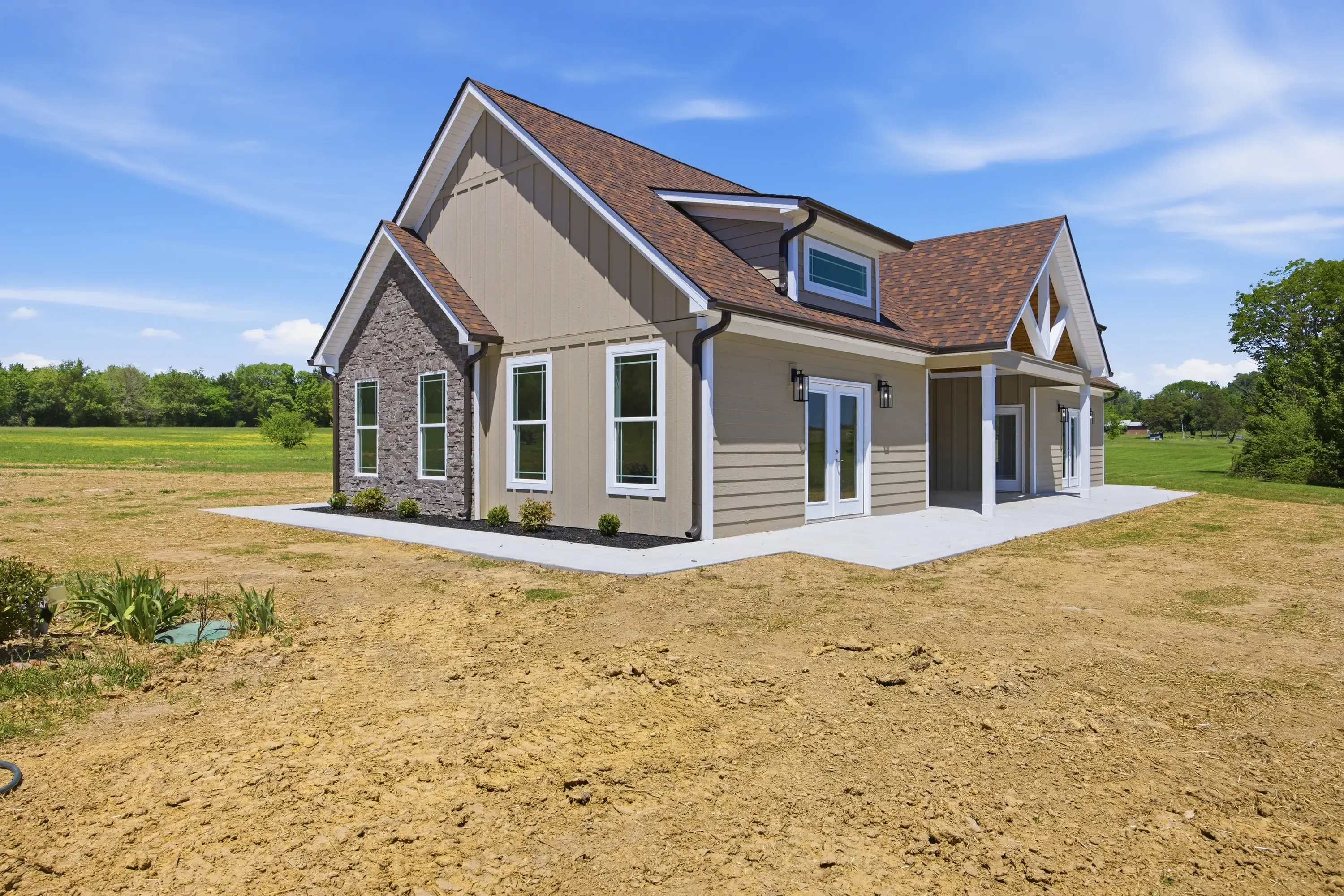 New farmhouse with stone veneer, tan siding, brown shingles, and concrete foundation on cleared lot